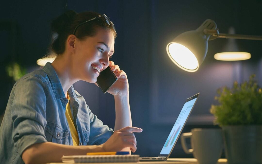 Smiling woman working at night and on a phone call while using her laptop